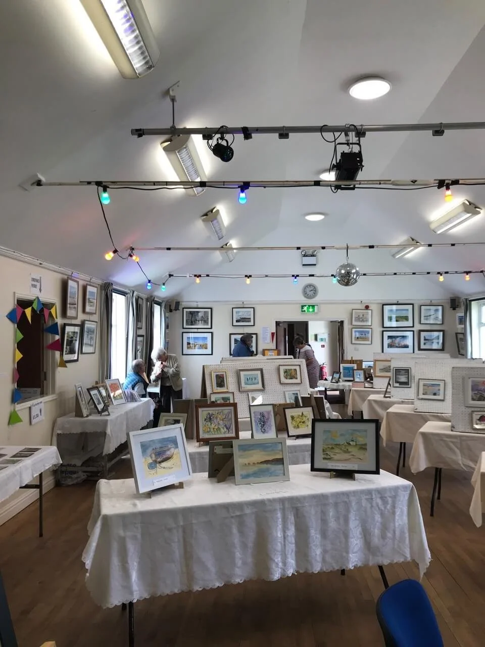 An art gallery with paintings and photographs on display, tables covered with white tablecloths holding framed artwork, and a few visitors viewing the art. The gallery has ceiling lights, a disco ball, and dark curtains on the windows. There are colorful string lights hanging from the ceiling.
