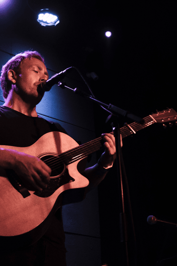 A man playing an acoustic guitar and singing into a microphone on a dark stage with purple and blue lighting.