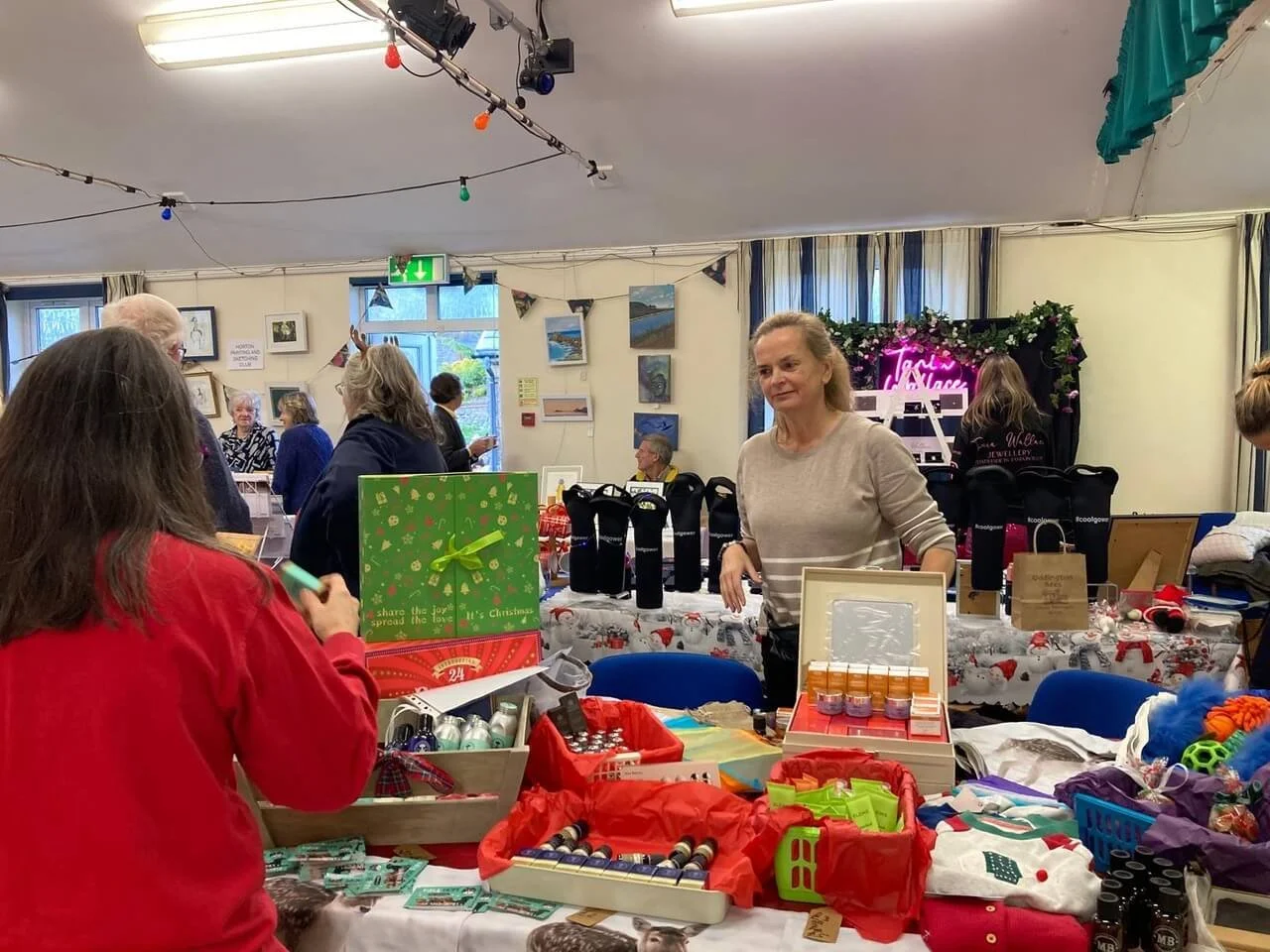 People shopping and browsing at a holiday market with tables full of gifts, decorations, and accessories. There are paintings on the walls and festive string lights hanging from the ceiling.