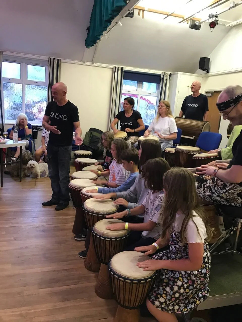 Children playing drums in a music class with adult instructors, some of whom are playing drums and others are supervising, in a room with windows, curtains, and a wooden floor.