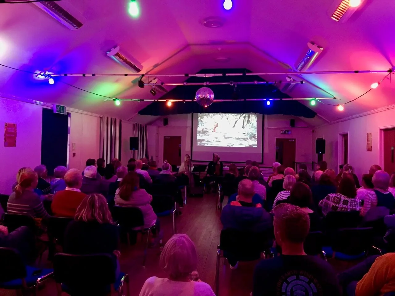 People seated in a dimly lit room watching a presentation on a large screen, with colorful stage lights, a disco ball, and audio equipment.