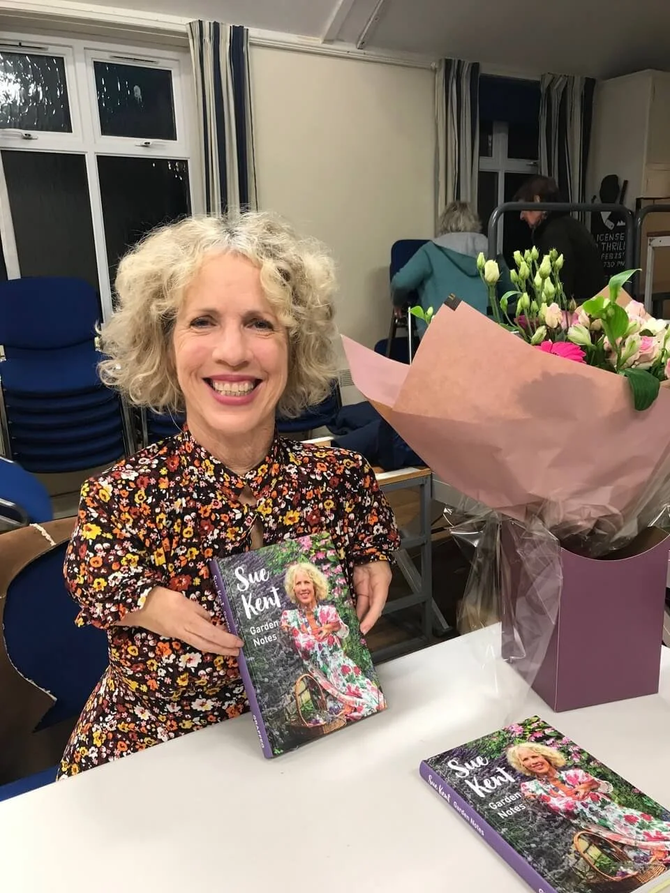 A woman with curly gray hair smiling and holding a book titled 'Sue Kent Garden Notes' at a table with a large bouquet of flowers. In the background, several people are seated near windows.