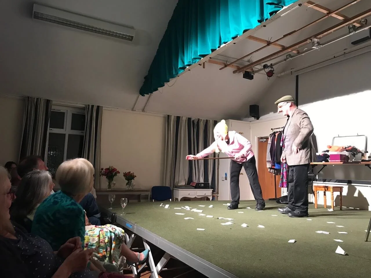 Two performers on stage entertaining a seated audience; one is bending down with a broom, and the other stands nearby watching, with props and costumes on a table to the side.