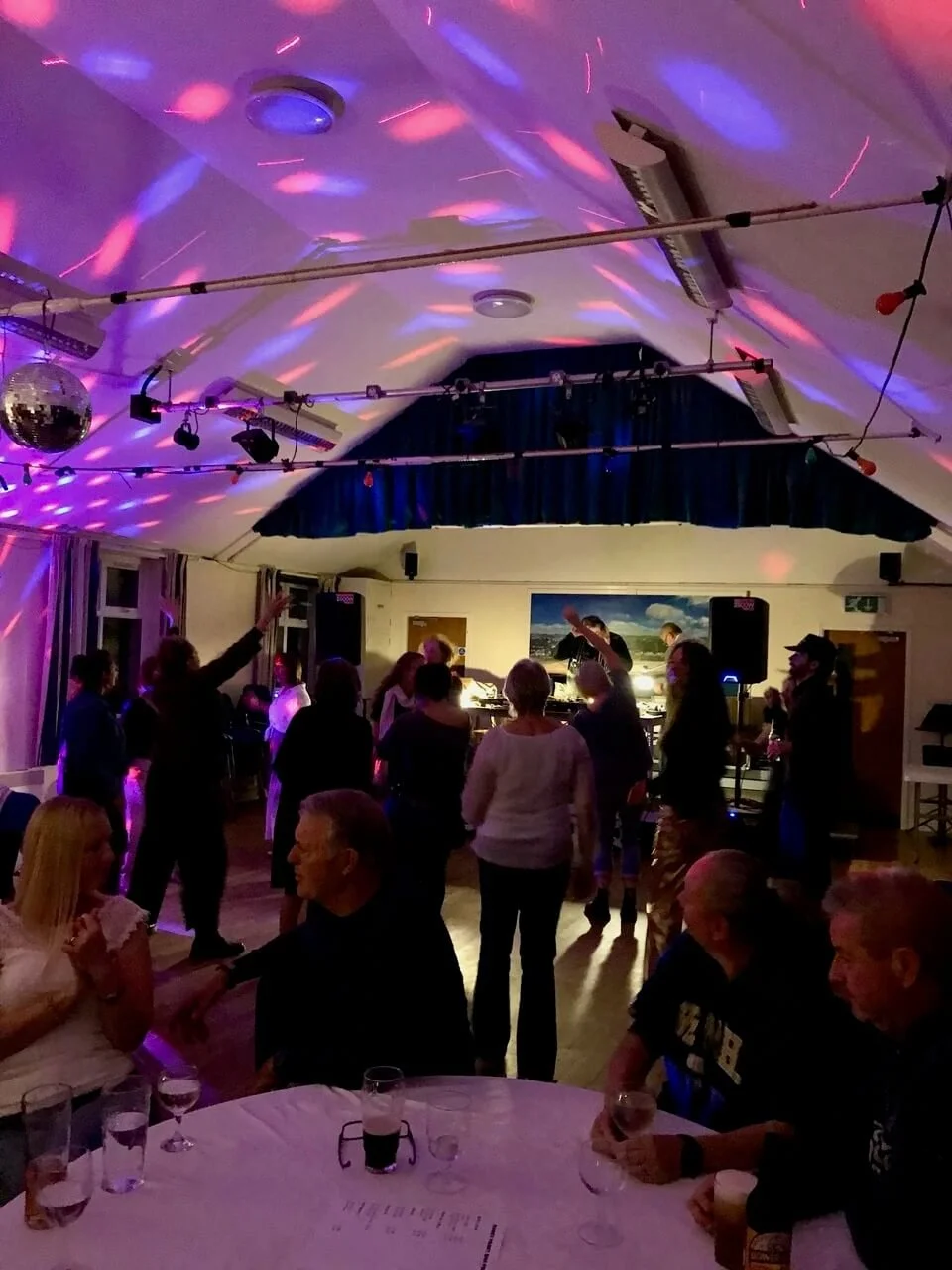 People dancing and socializing at a party with colorful lights and a DJ on stage in a decorated hall.