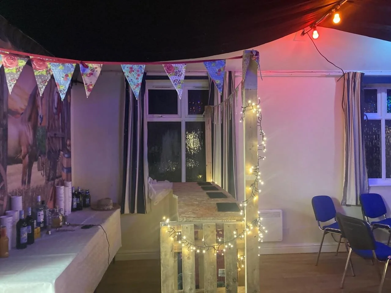 Indoor party setup with colorful bunting banners, string lights, a table with drinks, and chairs, illuminated by string and ceiling lights.