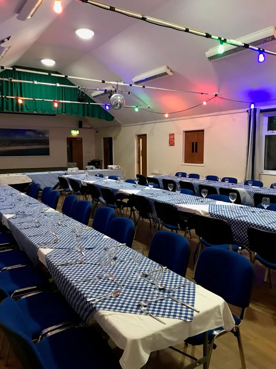 Event hall decorated with long tables covered with blue and white checkered tablecloths, set with glasses, forks, and spoons, ready for a meal. The ceiling has colorful string lights and a disco ball, creating a festive atmosphere. Walls are white wi