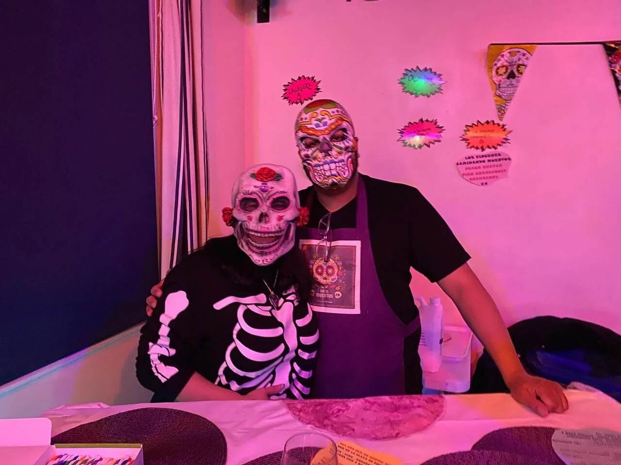 Two people wearing colorful skeleton masks pose in a festively decorated room with bright pink lighting, Día de los Muertos-themed decorations, and a table with a round centerpiece.
