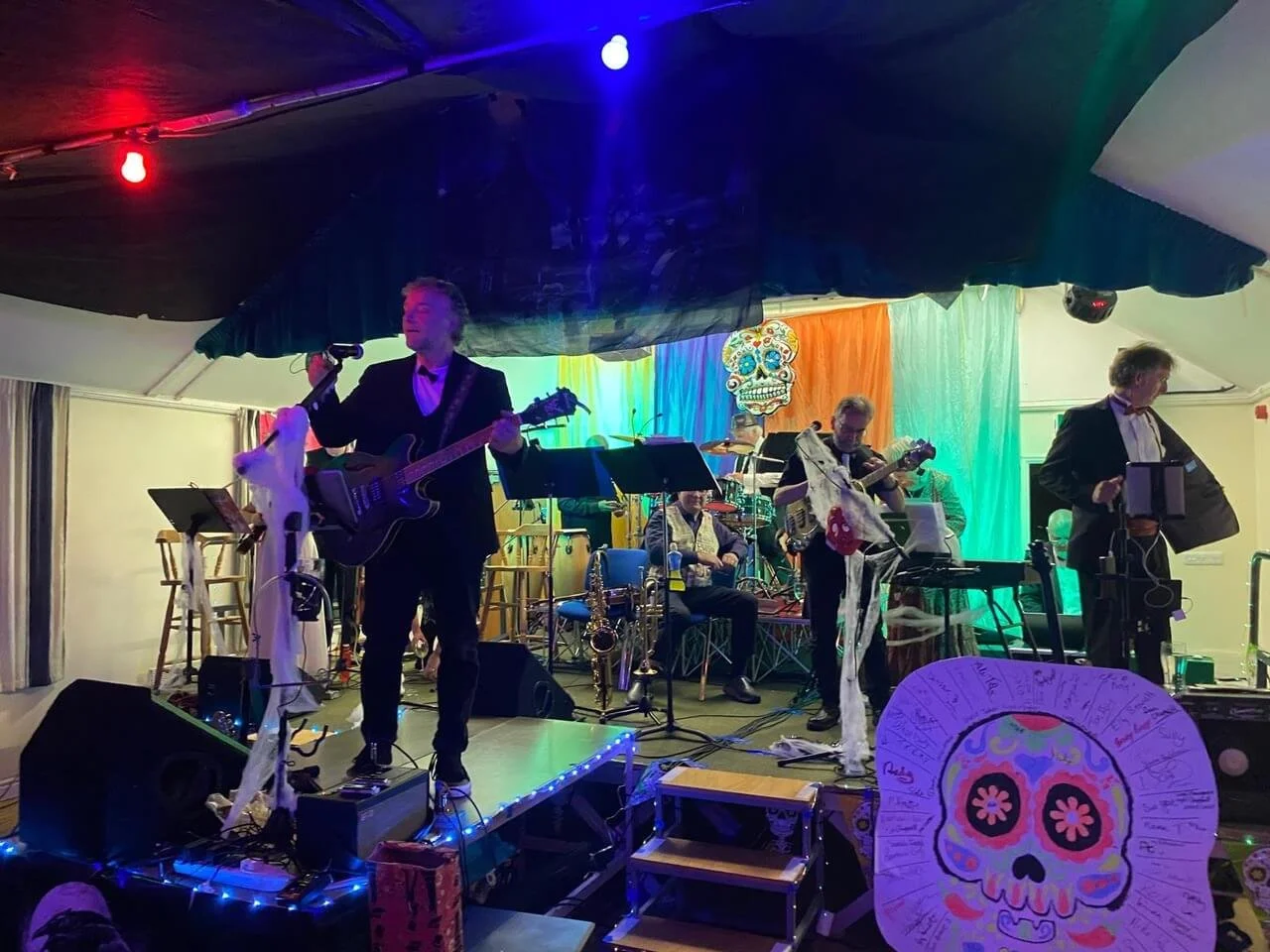 Band performing on stage with colorful backdrop including a decorative sugar skull.