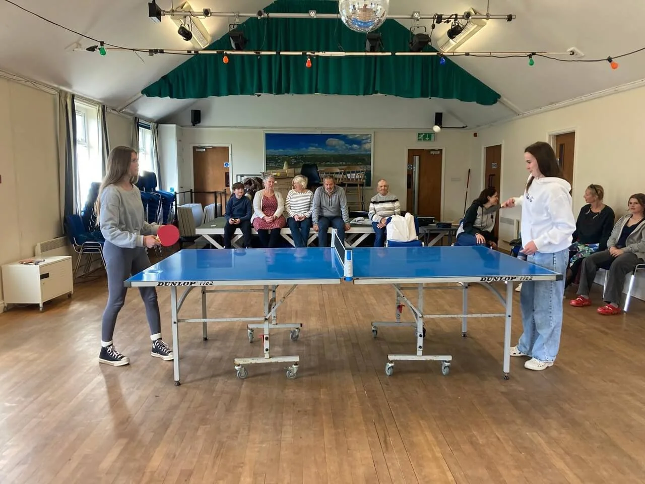 Two women playing table tennis indoors, with several spectators sitting along the back wall and watching the game.