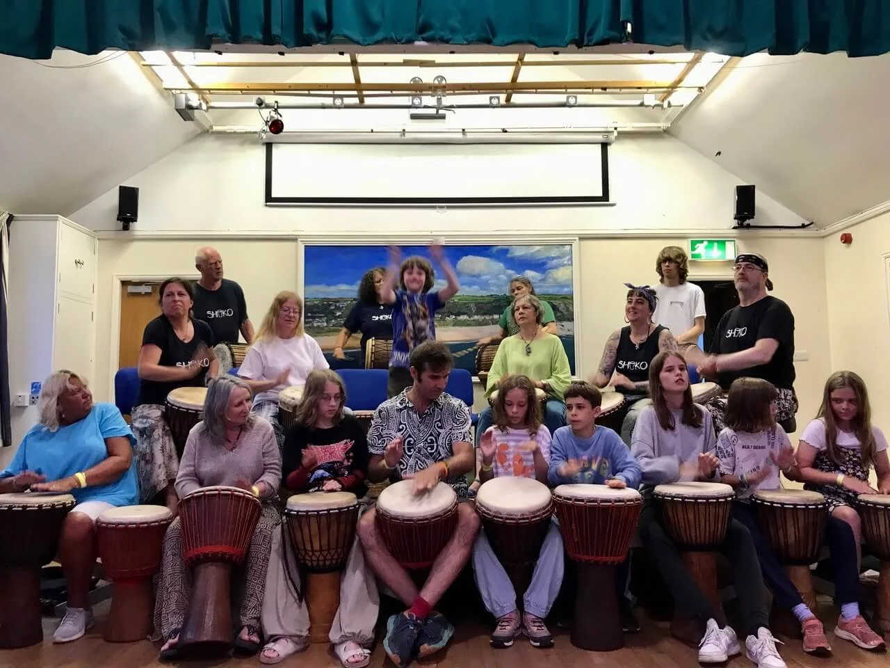 Group of children and adults playing drums in an indoor setting, with some standing and others seated, under a ceiling with skylights and a large painting on the back wall.