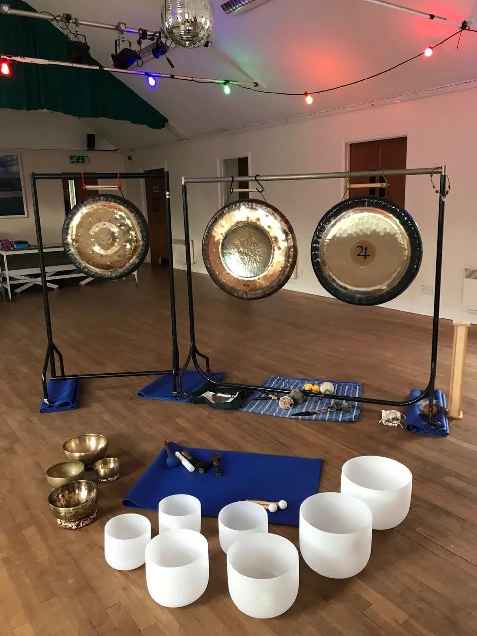 Three large gongs hanging on a frame, surrounded by various musical instruments including singing bowls and mallets, set up in a room with wooden floors, colorful string lights, and a disco ball hanging from the ceiling.