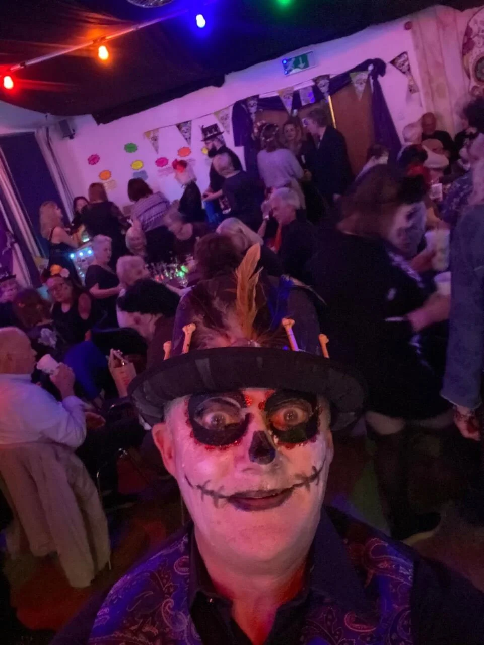 A person dressed as a skeleton with face paint, wearing a black hat with orange decorations, in a dimly lit party setting with colorful string lights and people socializing in the background.