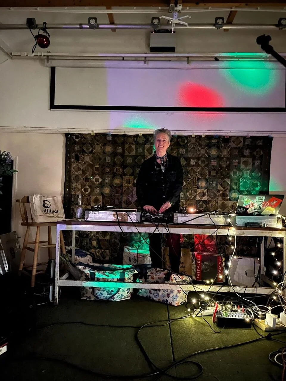 A woman standing behind a DJ console with colorful lights, in a cozy indoor venue with decorations and equipment.