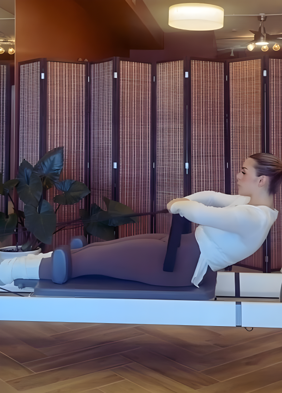 A woman in activewear on a reformer at Cat's Core Pilates studio with a wooden room divider and indoor plant in the background.