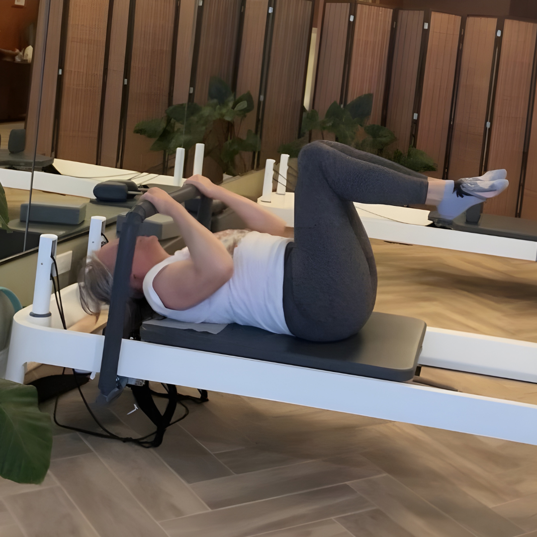 A woman is lying on her back on a Pilates reformer machine, holding onto the bars with her arms and lifting her legs with knees bent, in Cat's Core Pilates studio with a wooden wall and plants in the background.