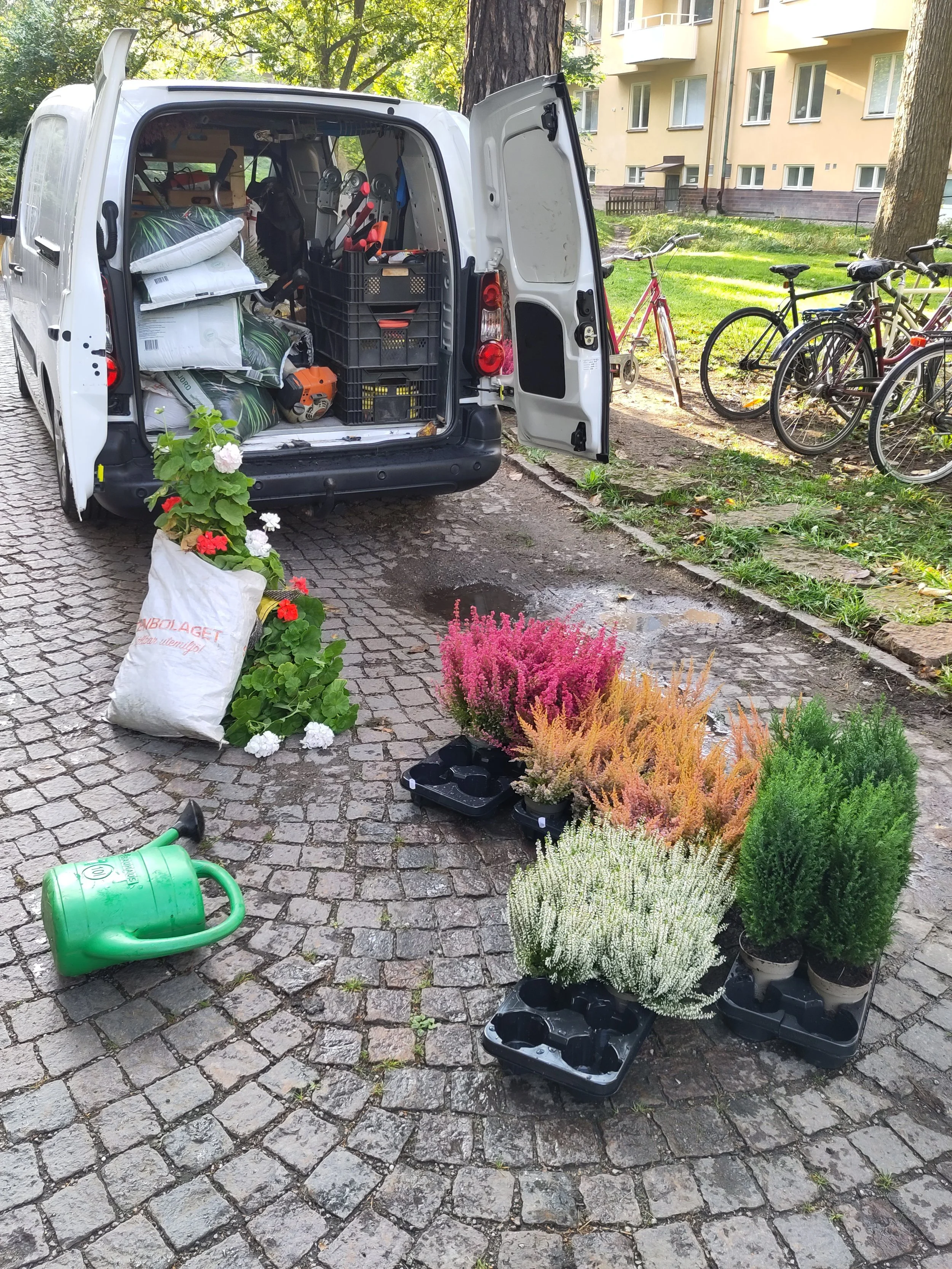 Open white van parked on a cobblestone path containing gardening tools, bags, and other supplies. Potted plants and trays of colorful flowering plants are on the ground nearby, with bicycles parked at a grassy area and multi-story residential building in the background.