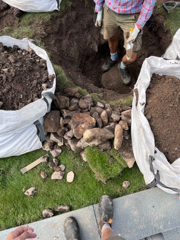 Person standing in a large hole using a yellow measuring tape, surrounded by white sandbags filled with soil or rocks, with rocks and grass around the hole, and another person taking the photo from a higher vantage point.