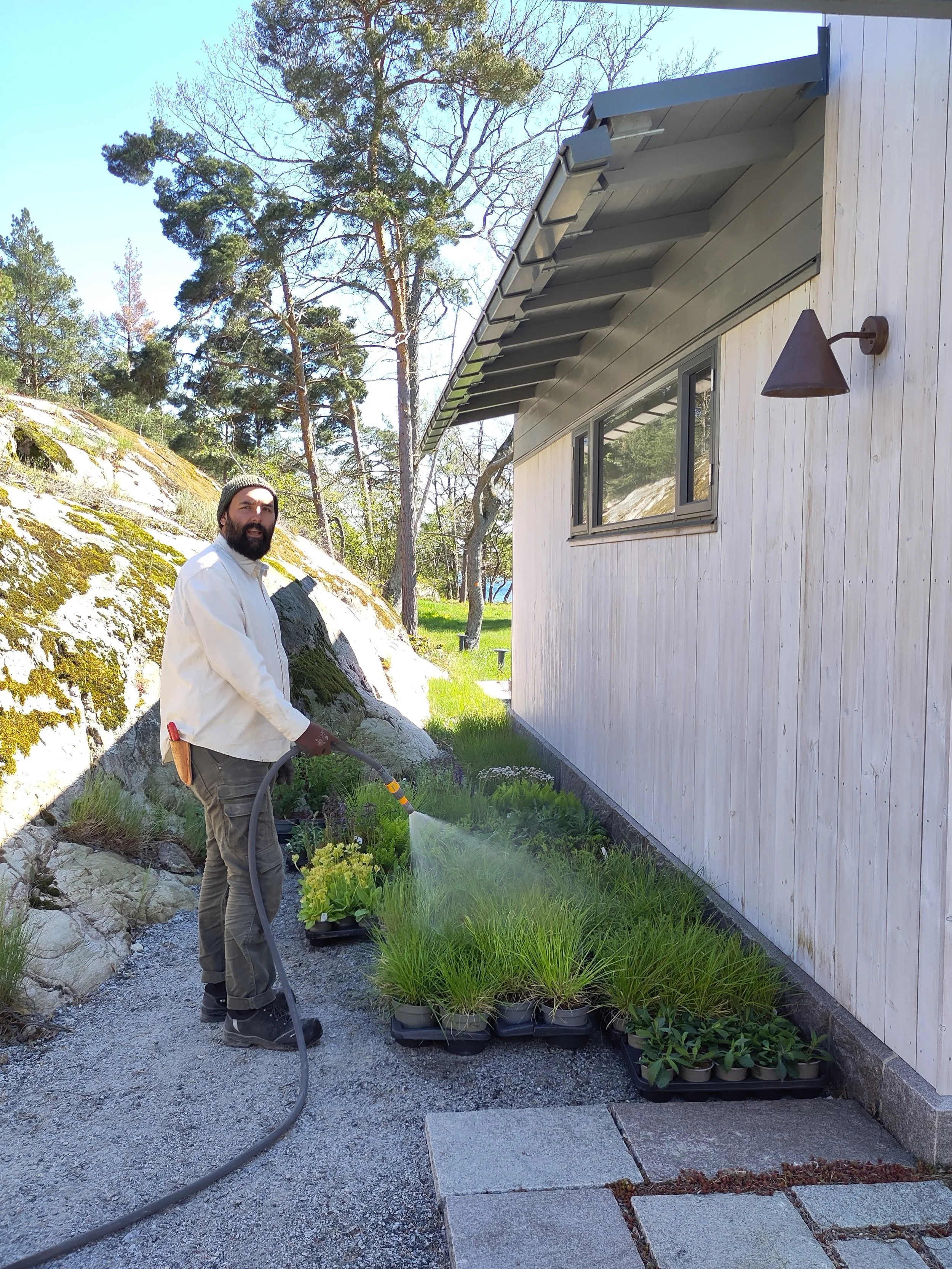 Man watering plants outside of a light-colored wooden house on a gravel path with trees in the background.