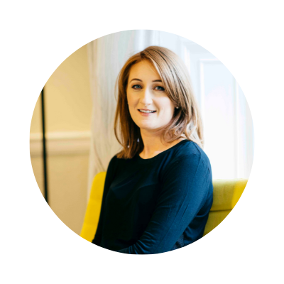 A woman with shoulder-length blonde hair, wearing a dark blue top, sitting on a yellow chair in a bright room with large windows and light-colored curtains.