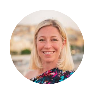 A smiling blonde woman outdoors with a cityscape background.