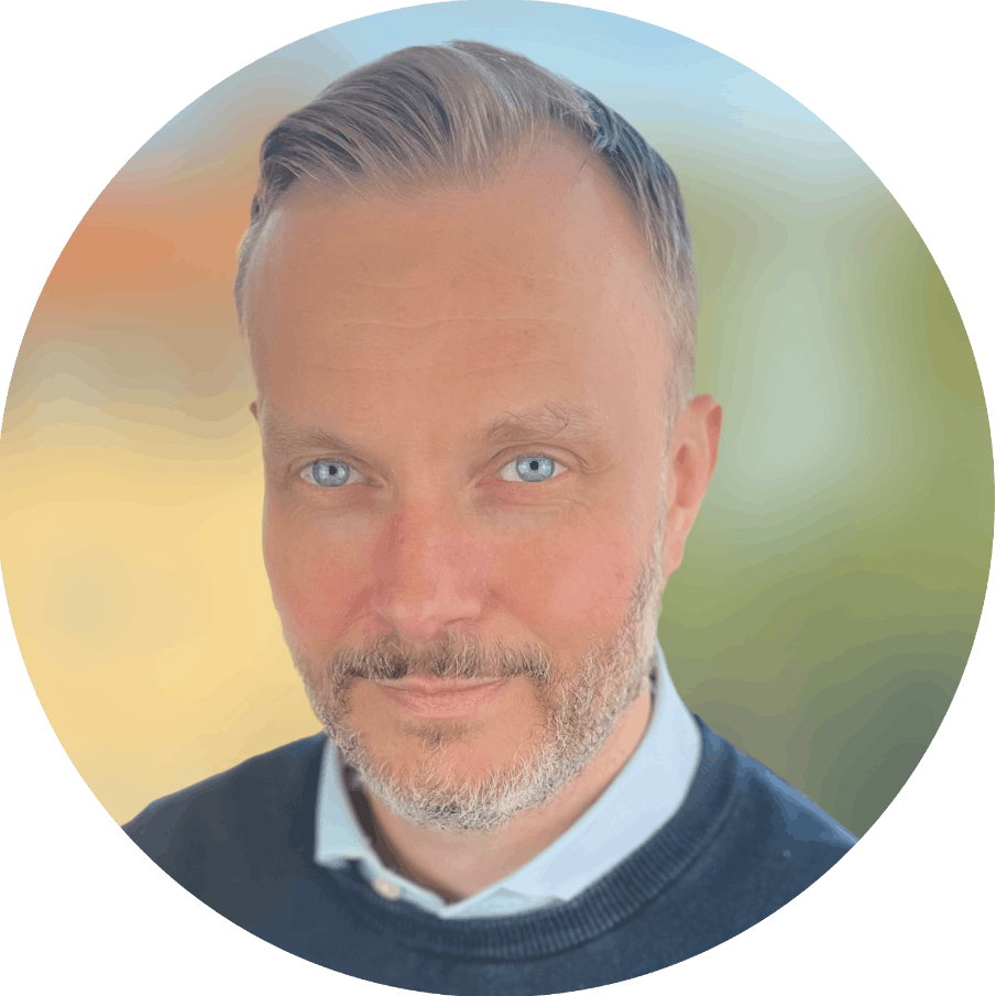 A middle-aged man with light skin, short gray hair, blue eyes, and a gray beard, wearing a dark sweater over a collared shirt, smiling softly against a blurred outdoor background.