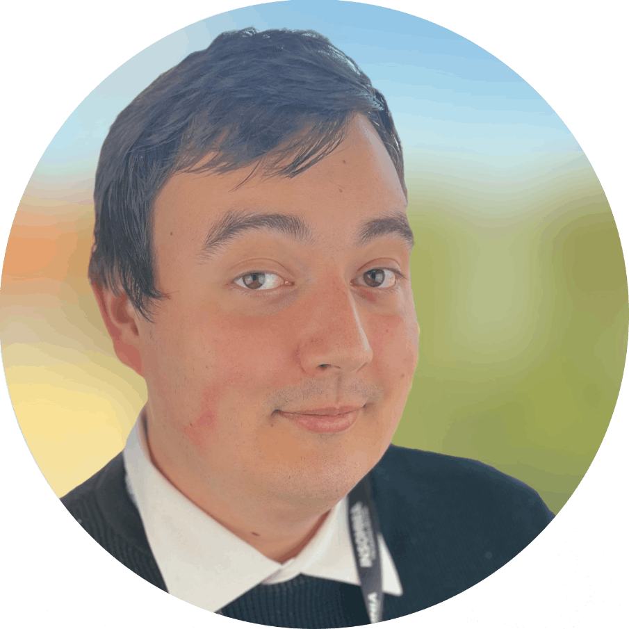 A young man with dark hair wearing a white shirt, black sweater, and lanyard, smiling softly with a blurred outdoor background.