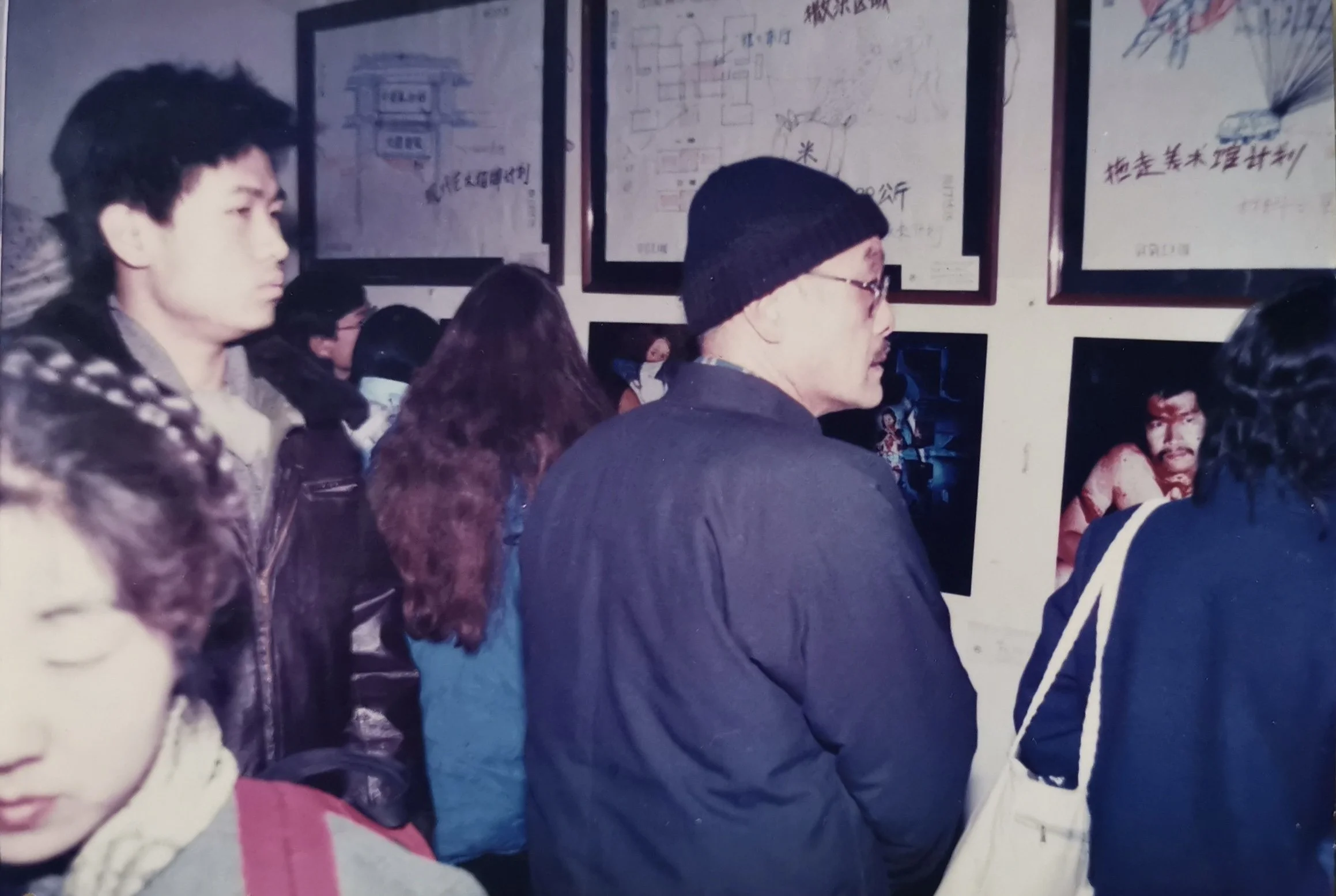 A group of people inside an art gallery, looking at framed art pieces on the wall. The people are of Asian descent, wearing casual clothing. Some are focused on the artwork, including a man with glasses and a beanie, and a woman with long hair.