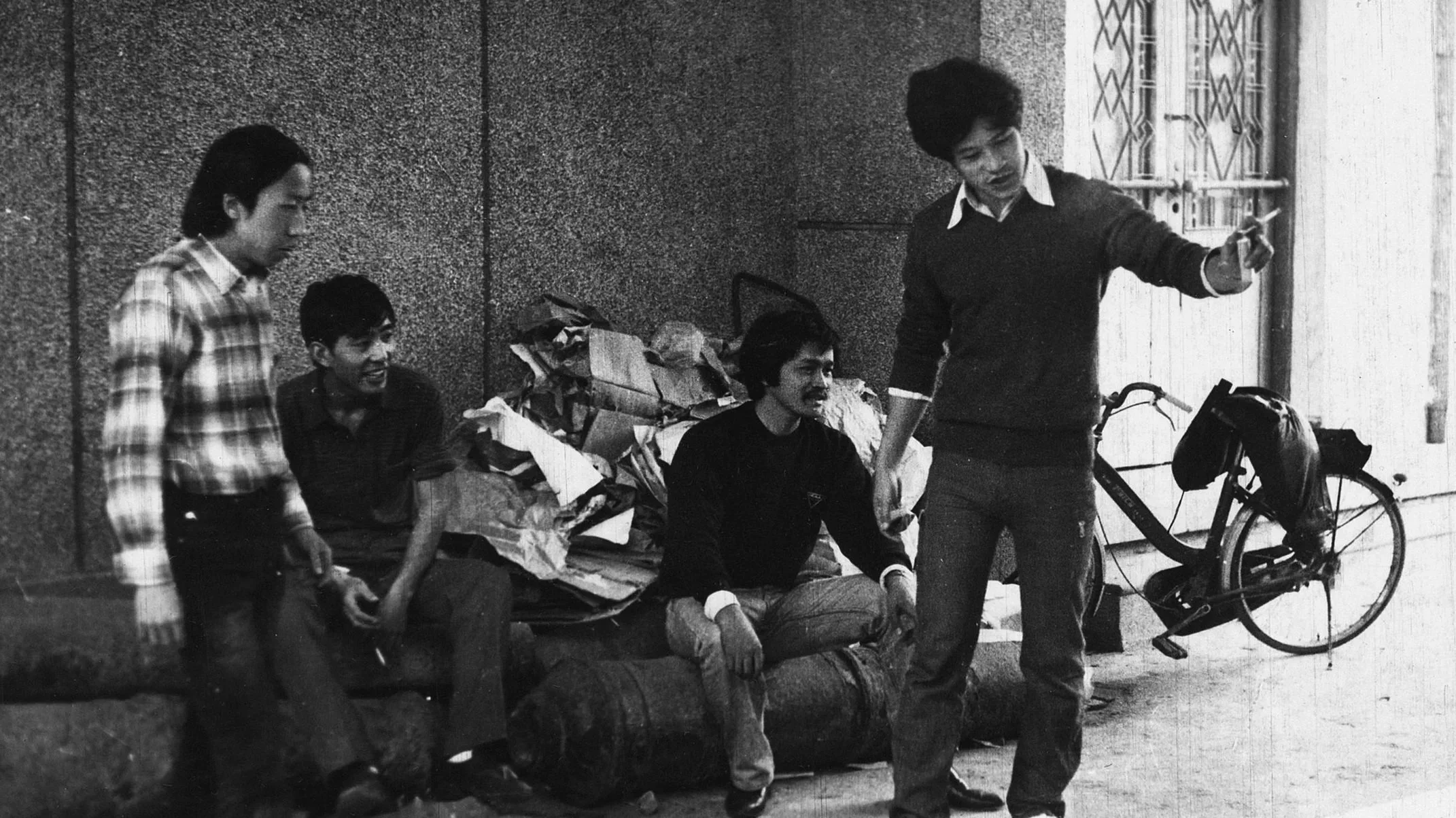 Four young men with dark hair and Asian features gather in an urban street, one of them taking a selfie with a smartphone, while the others sit and stand around a pile of trash and a bicycle is visible in the background.