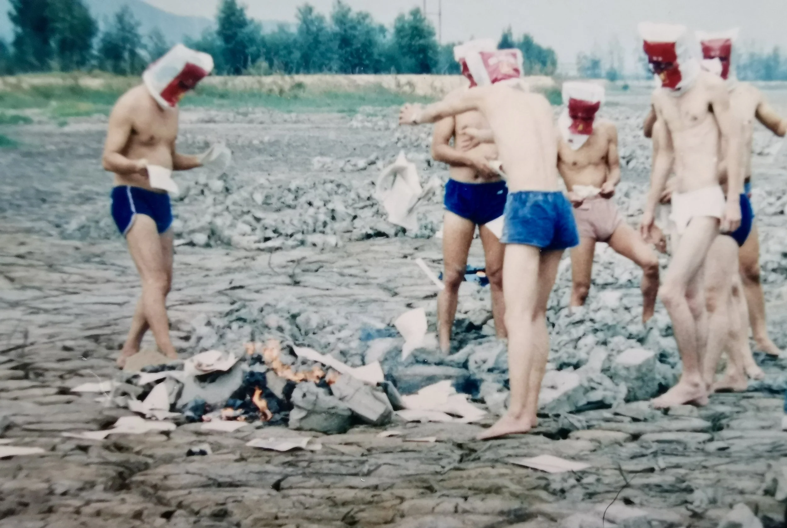 Seven shirtless men wearing swim trunks and red-and-white paper hats gathered around a small bonfire on rocky ground outdoors, with trees in the background.