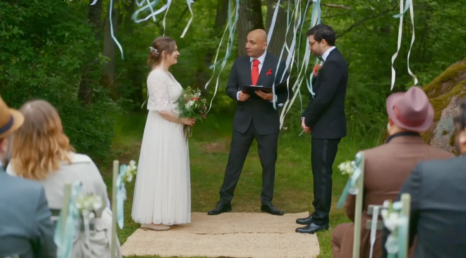 Par som gifter sig under en utomhusceremoni, med ett vitt brudklänning och en man i svart kostym, omgiven av vänner och släktingar i en grön skogsmiljö