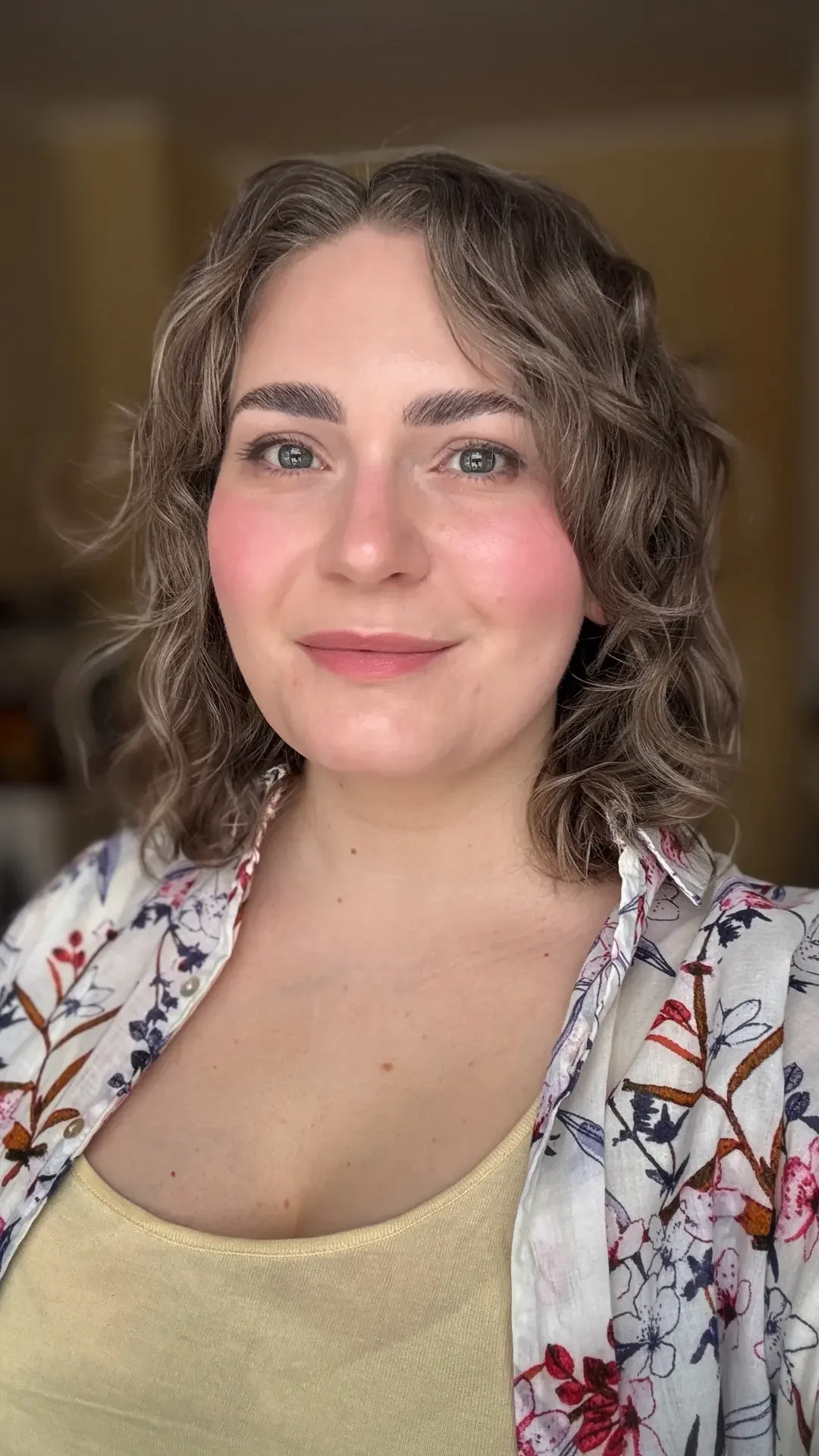 En ung kvinna med lockigt, brunt hår och ljus hy, som ler subtilt. Hon bär en ljusgul topp och en vit blommig skjorta. Bakgrunden är suddig med varma toner.