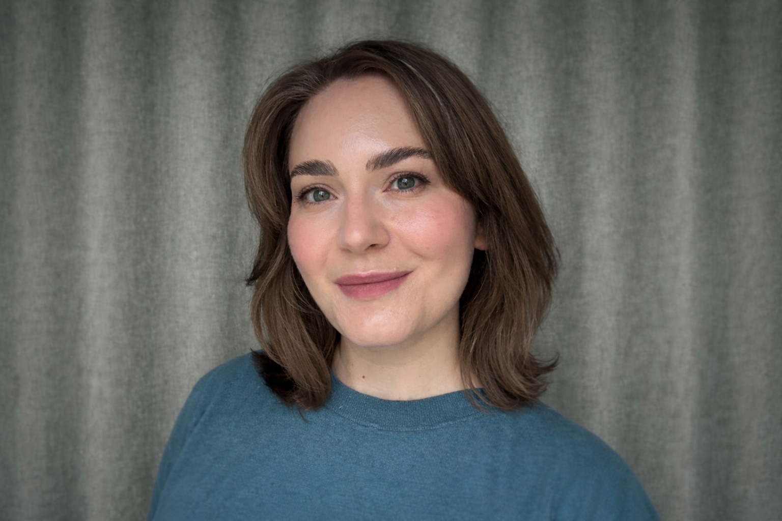 Porträtt av en ung kvinna med brunt axellångt hår, gröna ögon och giltig makeup. Hon ler svagt och bär en blå tröja framför en grå tygvägg.