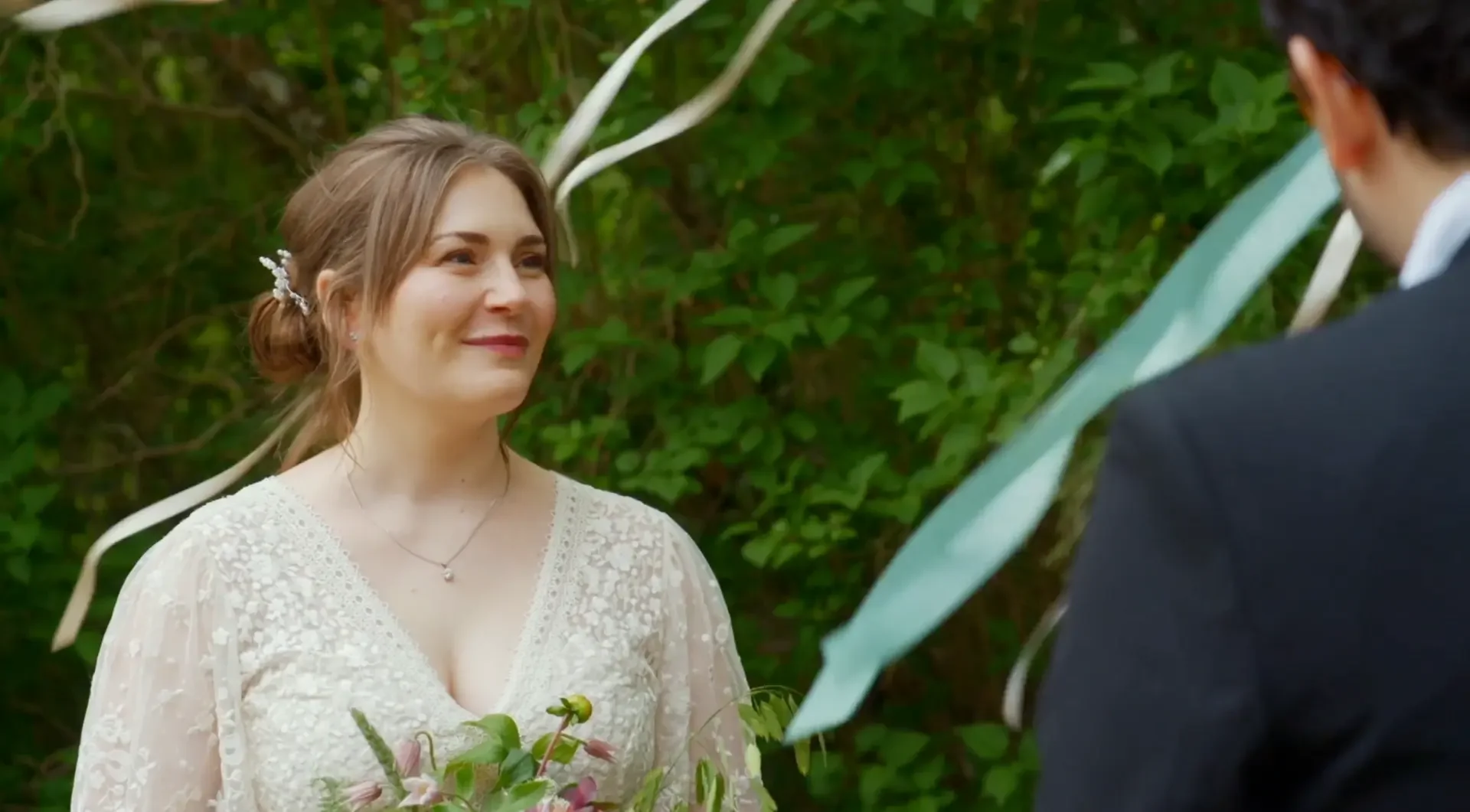 En brud och en man vid ett utomhusbröllop, bruden håller en bukett och ler emotionellt.