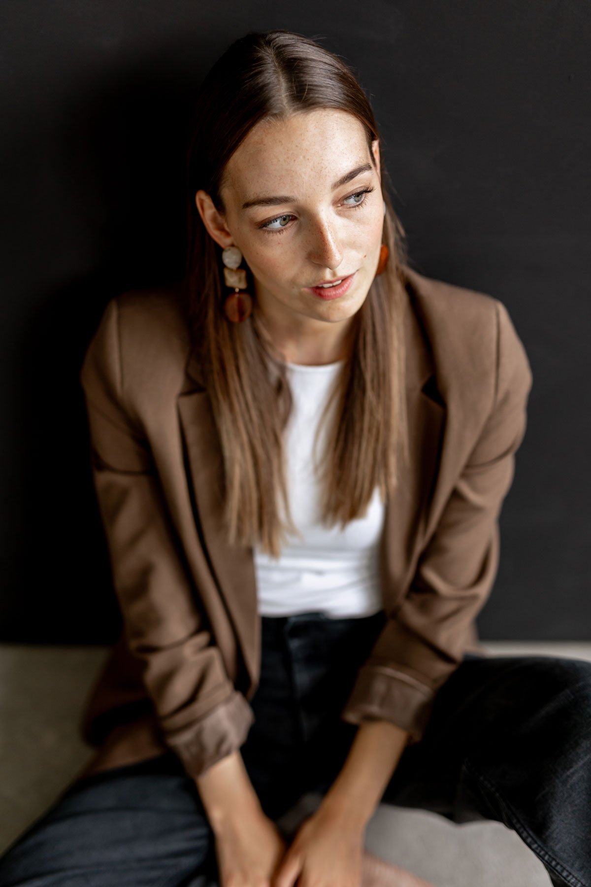 Junge Frau mit braunen Haaren, brauner Jacke, weißen T-Shirt und schwarzen Hosen sitzt auf dem Boden vor einem schwarzen Hintergrund.