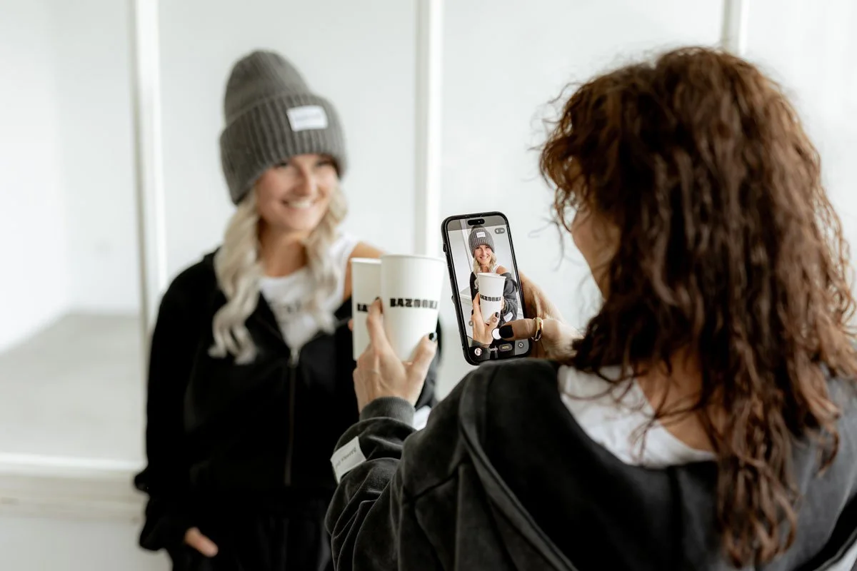 Zwei Frauen bei einem Fotoshooting, eine hält eine Tasse, während die andere ein Smartphone ausrichtet, um ein Bild zu machen.