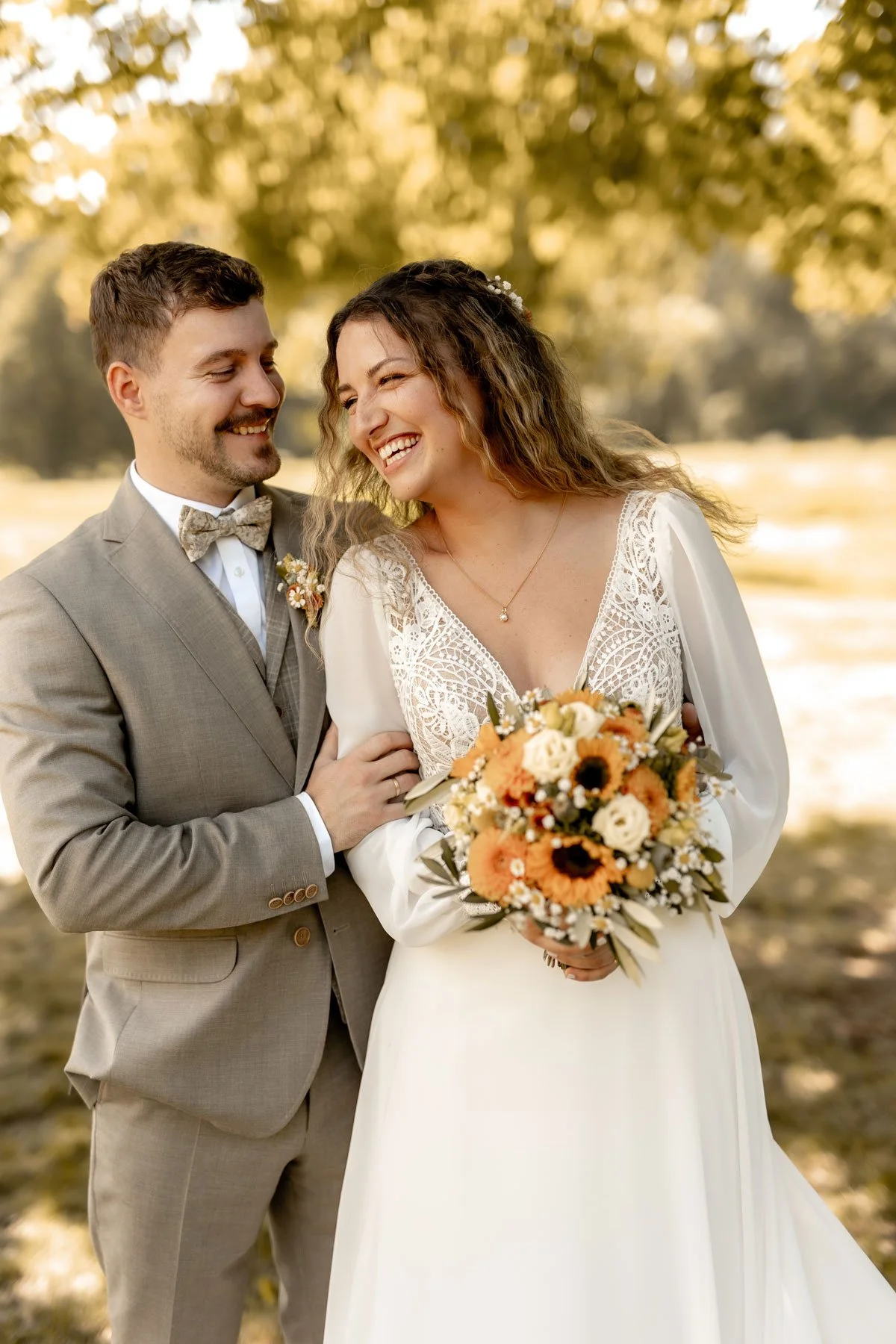Ein Paar bei einer Hochzeit im Freien, die Braut hält einen Blumenstrauß und beide lachen in die Kamera.