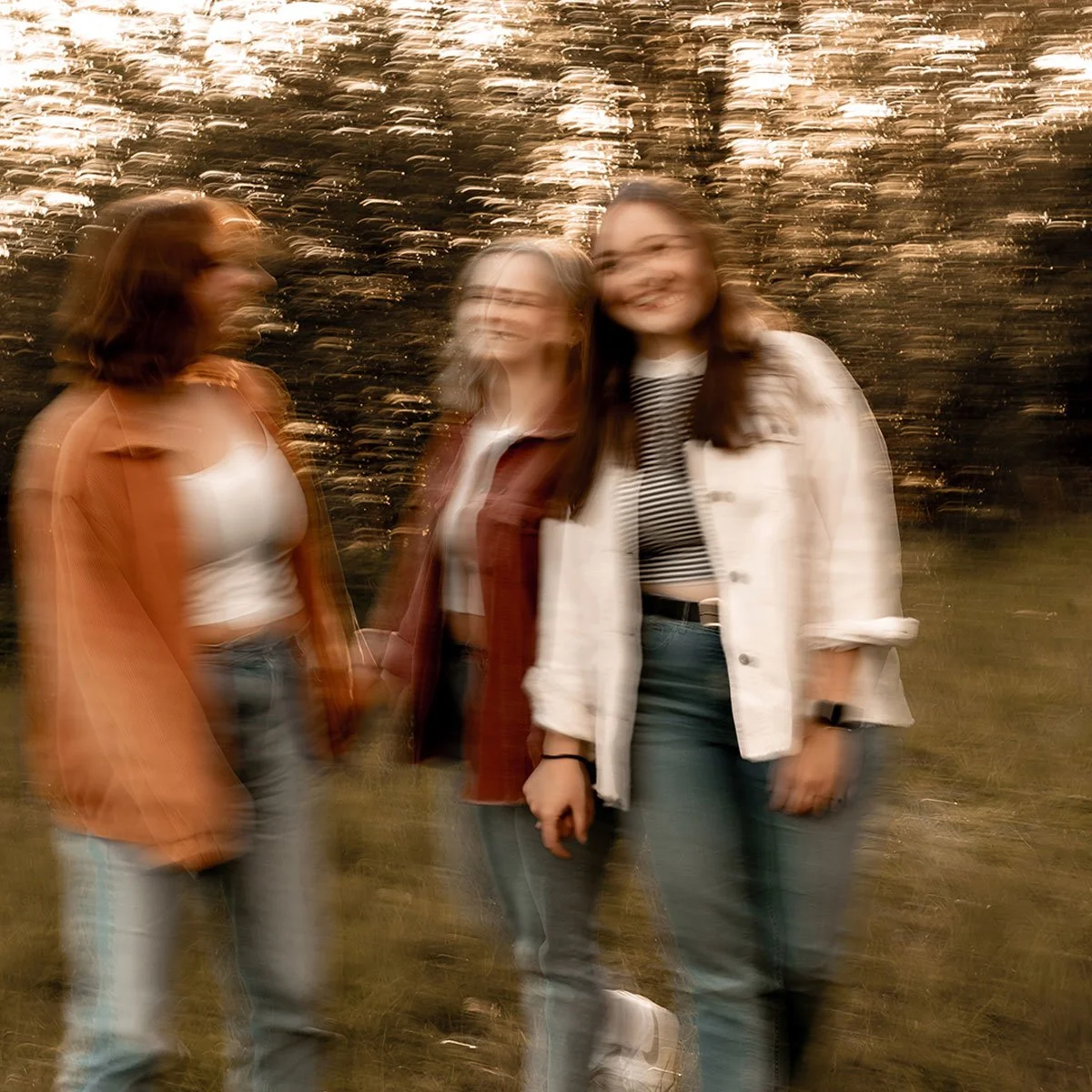 Drei lachende Frauen stehen im Freien bei Sonnenuntergang, der Himmel ist golden, die Bewegung verursacht Unschärfe im Bild.