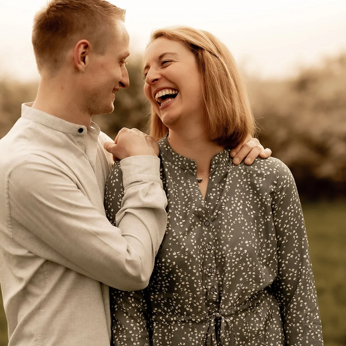 Ein lachendes Paar, das sich im Freien nahe beieinander umarmt, im Hintergrund nebliges Herbstlaub.