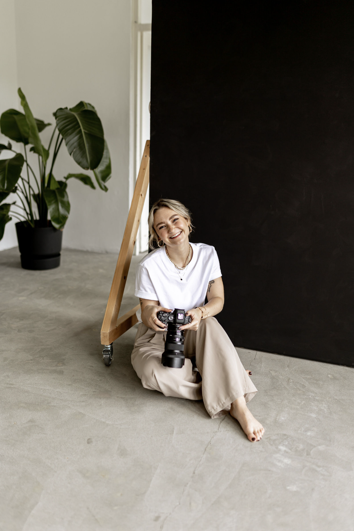 Eine lachende Frau sitzt auf dem Boden in einem Fotostudio, hält eine Kamera und ist neben einem schwarzen Hintergrund und einer großen Pflanzen. Sie trägt ein weißes T-Shirt und beige Hosen.