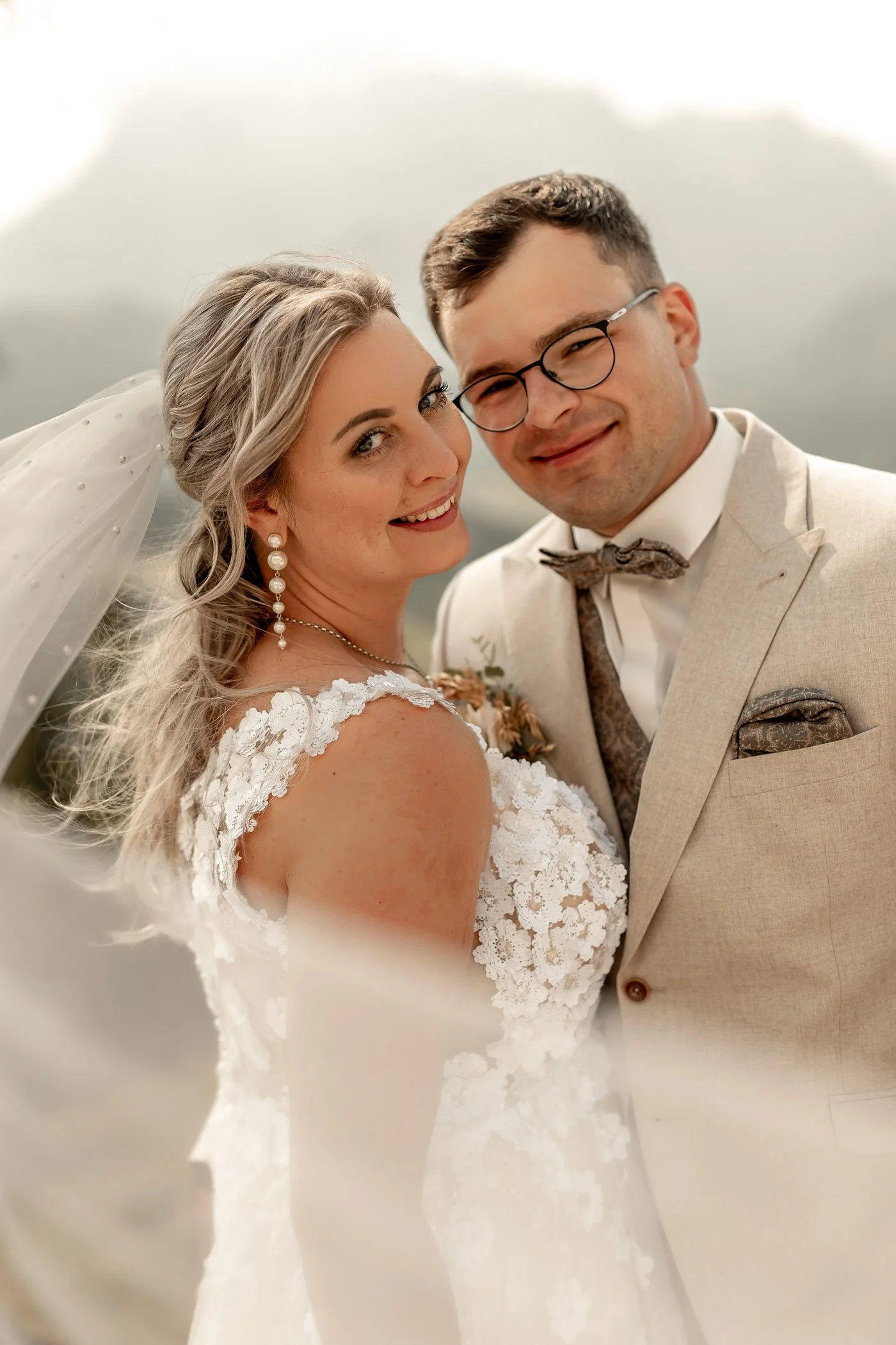 Ein frisch verheiratetes Paar bei ihrer Hochzeit, lachend und in eleganter Kleidung, im Freien mit bewölktem Himmel im Hintergrund.