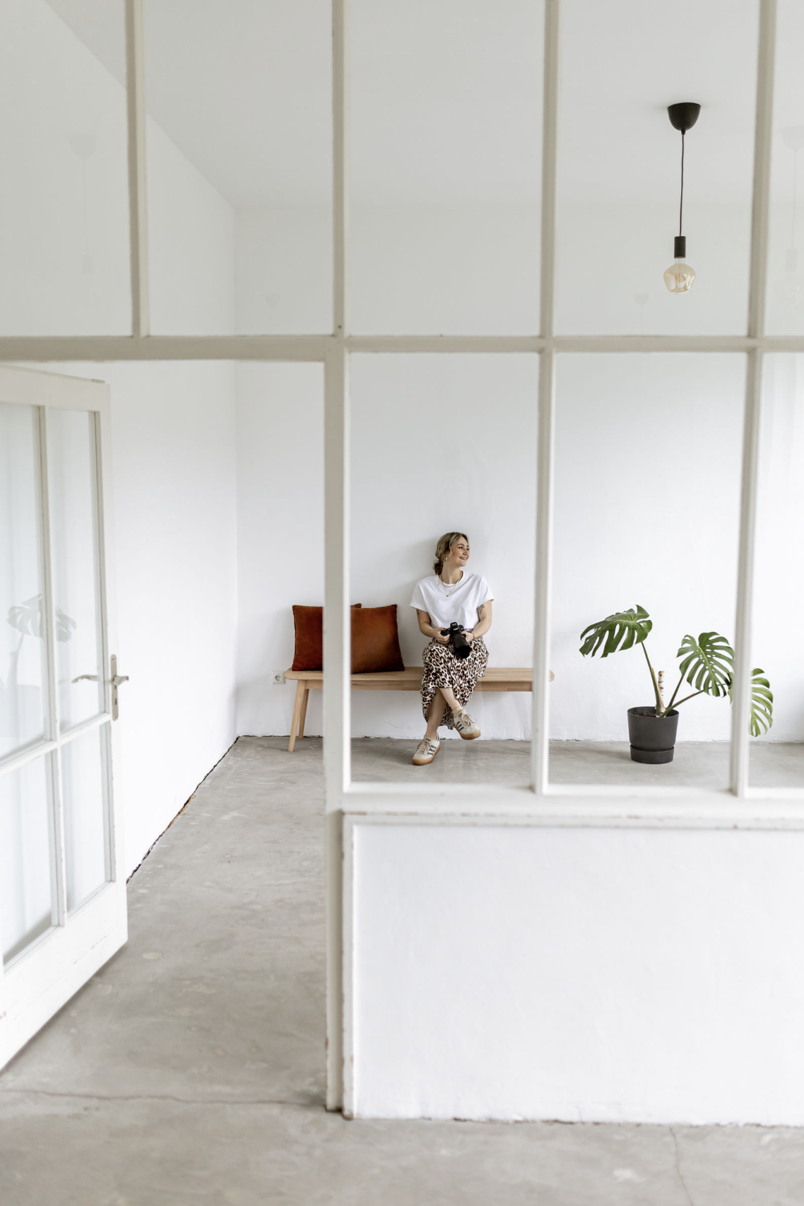 Eine junge Frau sitzt auf einer Holzbank in einem weißen Raum, hält eine Kamera in den Händen und lächelt. Es gibt zwei braune Kissen auf der Bank und eine große Topfpflanze neben ihr.