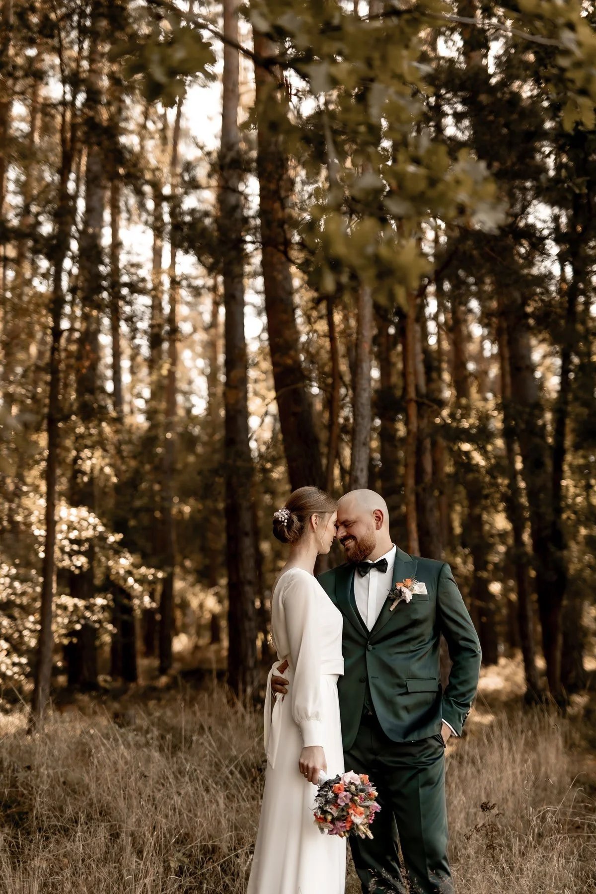 Ein Brautpaar steht im Wald, eng aneinander, lächelnd und mit der Stirn aneinander. Die Braut trägt ein langärmeliges weißes Hochzeitskleid und hält einen Blumenstrauß, der Bräutigam trägt einen grünen Anzug mit Fliege und hat einen Blumenanstecker. 
