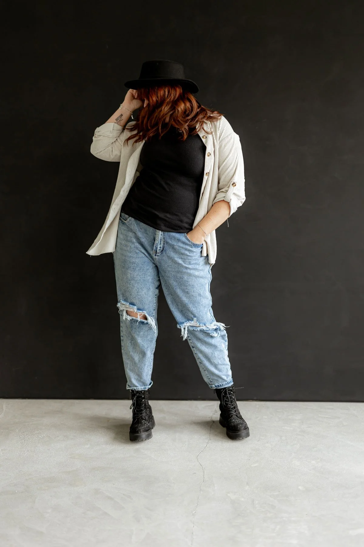 Frau in lässiger Kleidung mit schwarzem Hut, schwarzen Stiefeln, schwarzen T-Shirt, weißer Jacke und zerrissenen Jeans vor schwarzem Hintergrund.