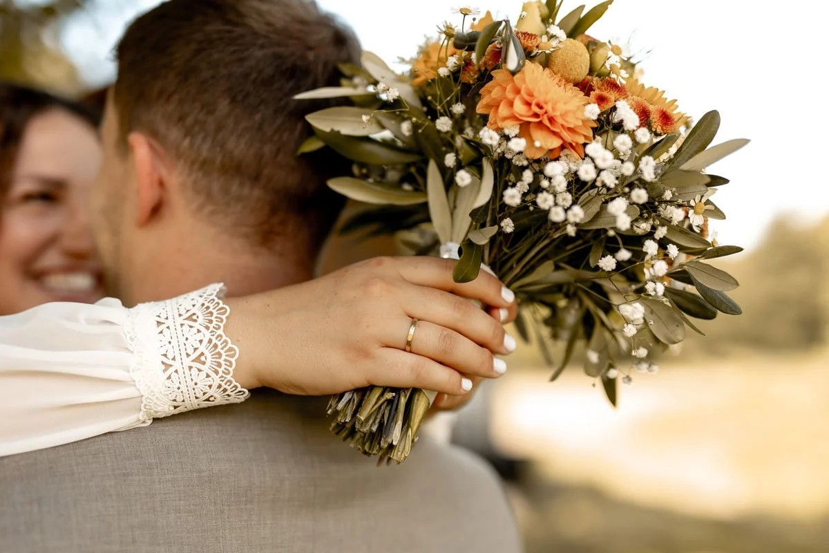 Nahaufnahme eines Brautpaares, wobei die Braut einen Blumenstrauß hält und die Hand mit Ehering auf den Rücken des Bräutigams legt.