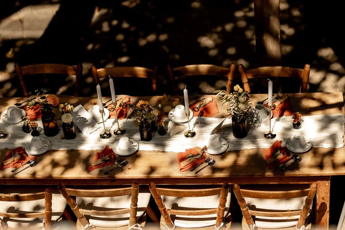 Ein festlich gedeckter Holztisch mit Kerzen, Blumenarrangements, Weißes Geschirr und Besteck, dekorative Stoffservietten, umgeben von Holzstühlen im Freien bei Sonnenlicht.