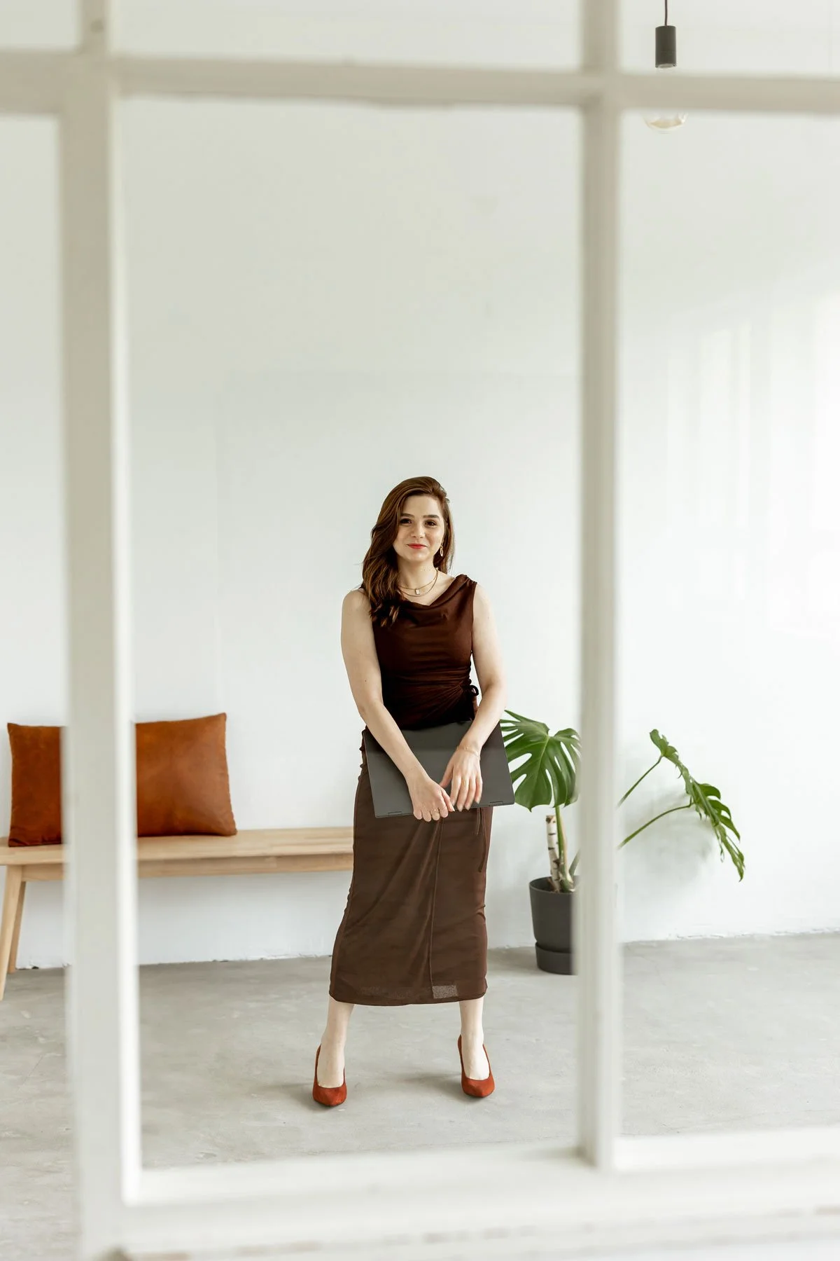 Eine junge Frau steht in einem minimalistischen Raum mit weißen Wänden, gehaltenem dunkelbraunem Kleid, roten Schuhen und einer schwarzen Mappe, im Hintergrund sind eine Holzbank mit zwei braunen Kissen und zwei grüne Topfpflanzen sichtbar.
