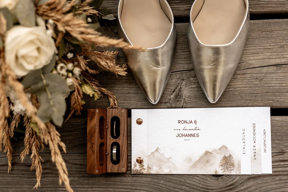 Hochzeits-Einladungskarte mit Bergmotiv, silberne Schuhe, eine Holzzahl, Blumen und Trockenpflanzen auf einem Holztisch.