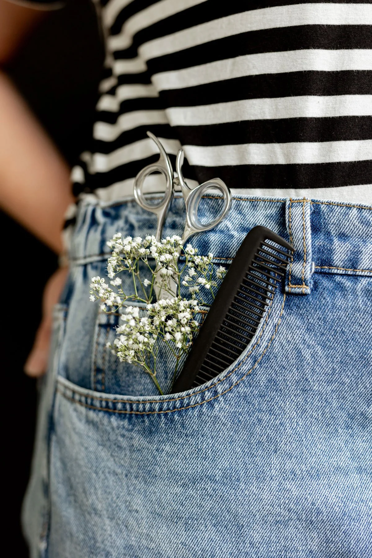 Ein Jeans-Tascheninhalt mit einer schwarzen Kamm, einer Schere und kleinen weißen Blumen, getragen von einer Person in einer schwarz-weißen gestreiften Bluse.