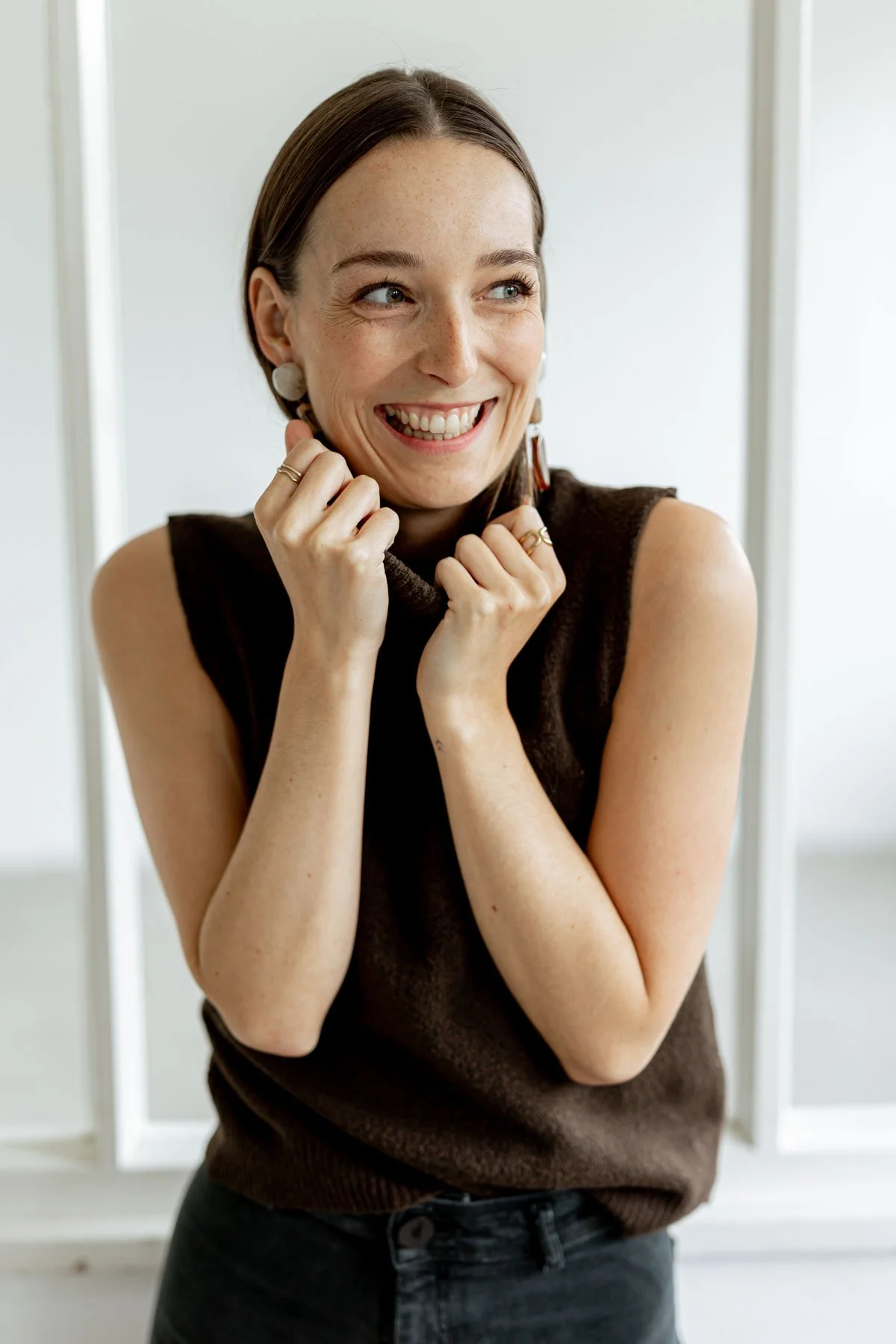 Eine lachende Frau mit braunen Haaren, die in einem dunklen Weste und schwarzen Jeans gekleidet ist, steht vor einer weißen Wand. Sie sieht glücklich aus.