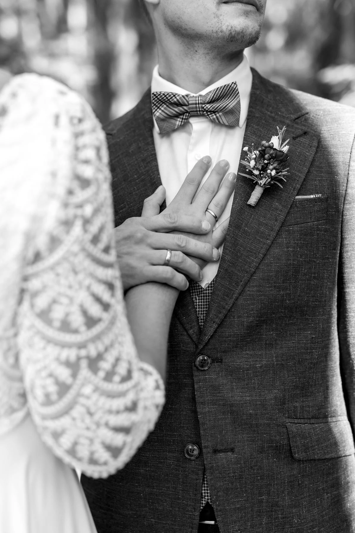 Nahaufnahme eines Paares bei ihrer Hochzeit, der Mann trägt einen Anzug mit Fliege und einen Anstecker, die Hand der Braut mit Ehering liegt auf seiner Brust, im Hintergrund unscharfe Natur.